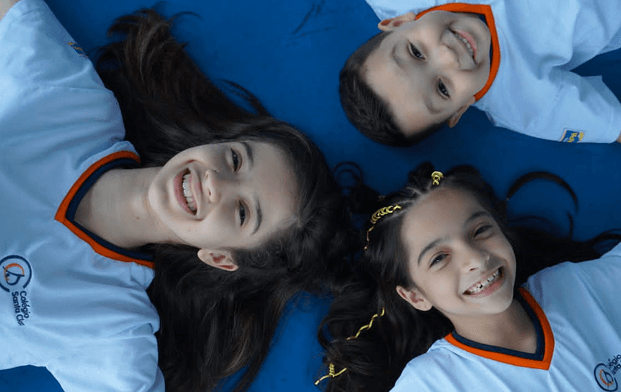Três crianças sorrindo deitadas em círculo no chão azul, usando uniformes escolares do Colégio Santa Clara na cor branca com gola laranja e azul.